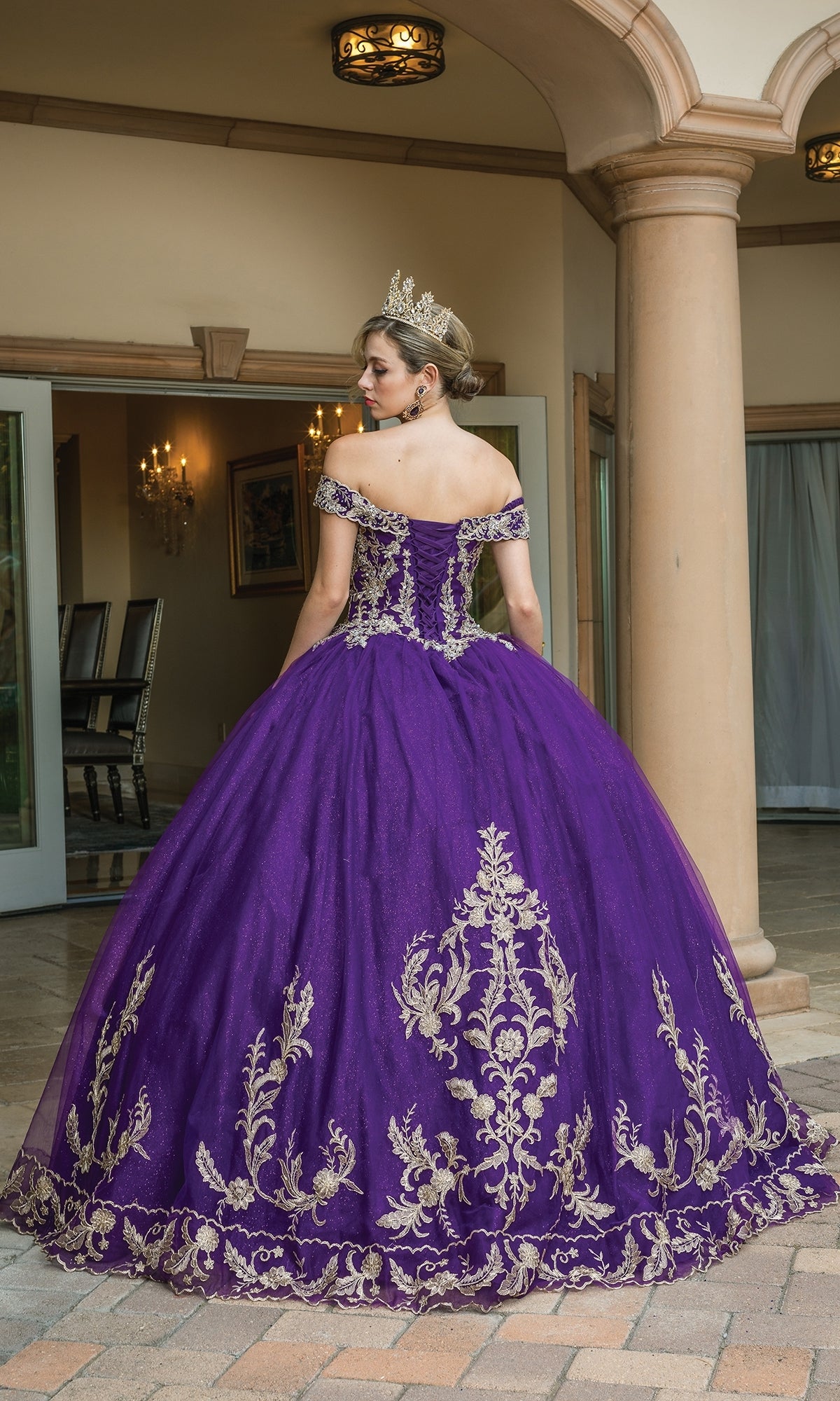 A woman in the Quinceanera Dress 1596 by Dancing Queen, featuring a gold-embroidered a-line ball gown and crown, stands outside a columned building with her back to the camera.